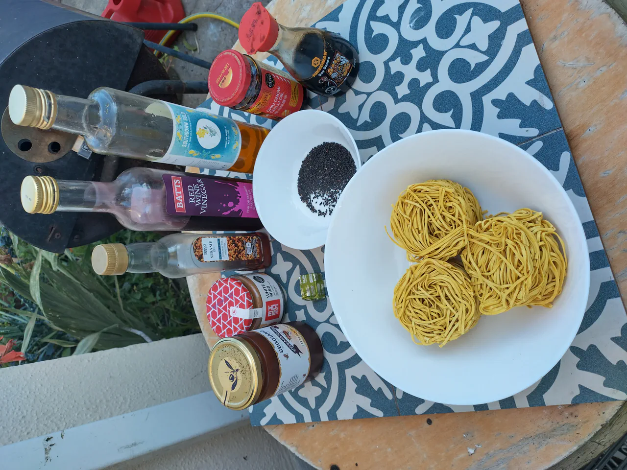 Egg noodle nests on a white plate alongside bottles of soy sauce, sesame oil, rice wine vinegar, chilli sauce, and a small bowl of black sesame seeds
